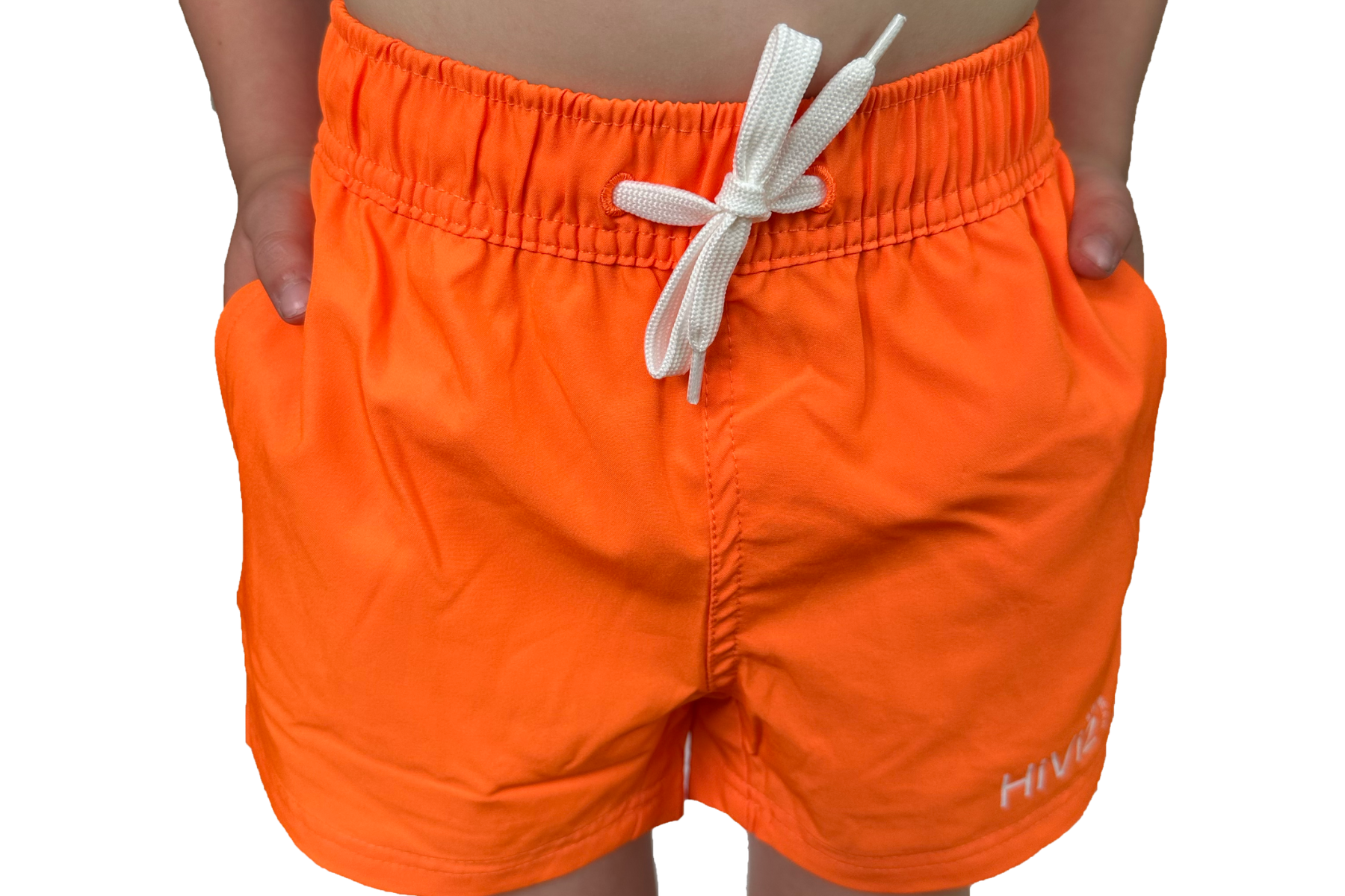 Toddler wears bright neon orange swim shorts with his hands in his pockets and the white drawstring in a bow.